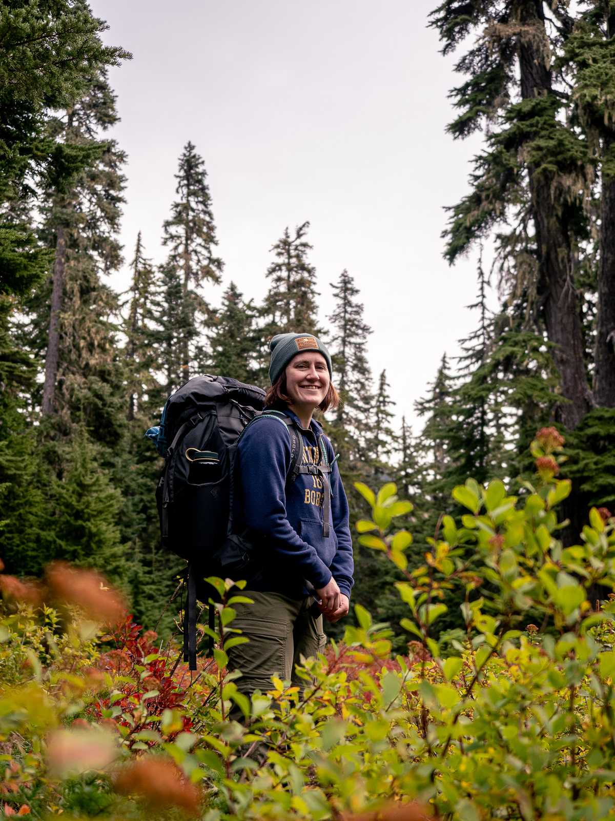 A female-presenting hiker smiles with pine trees in the background and green bushes in the foreground