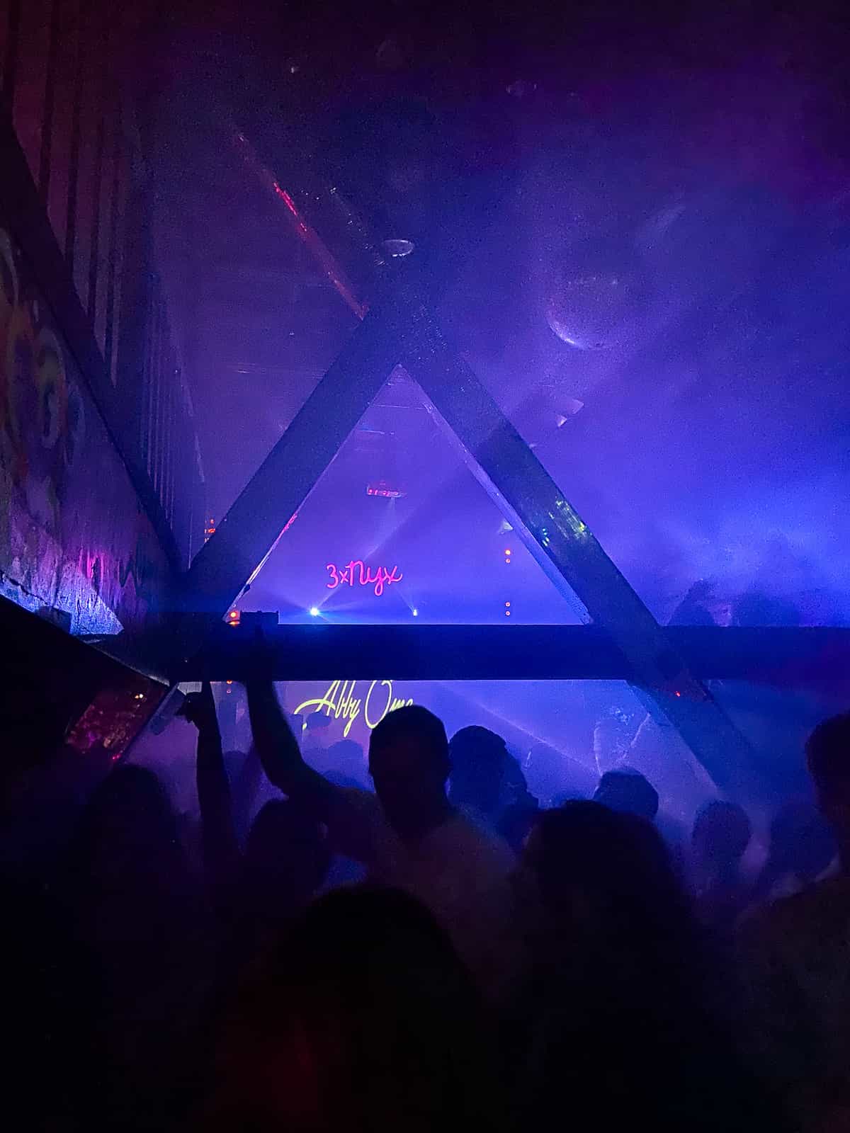 Purple and pink hues shine through the fog of a nightclub, with a disco ball hanging from the ceiling. A neon sign says "3xNYX"