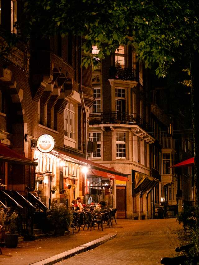 A warmly lit street at nighttime in Amsterdam, with people in outdoor seating outside a bar.