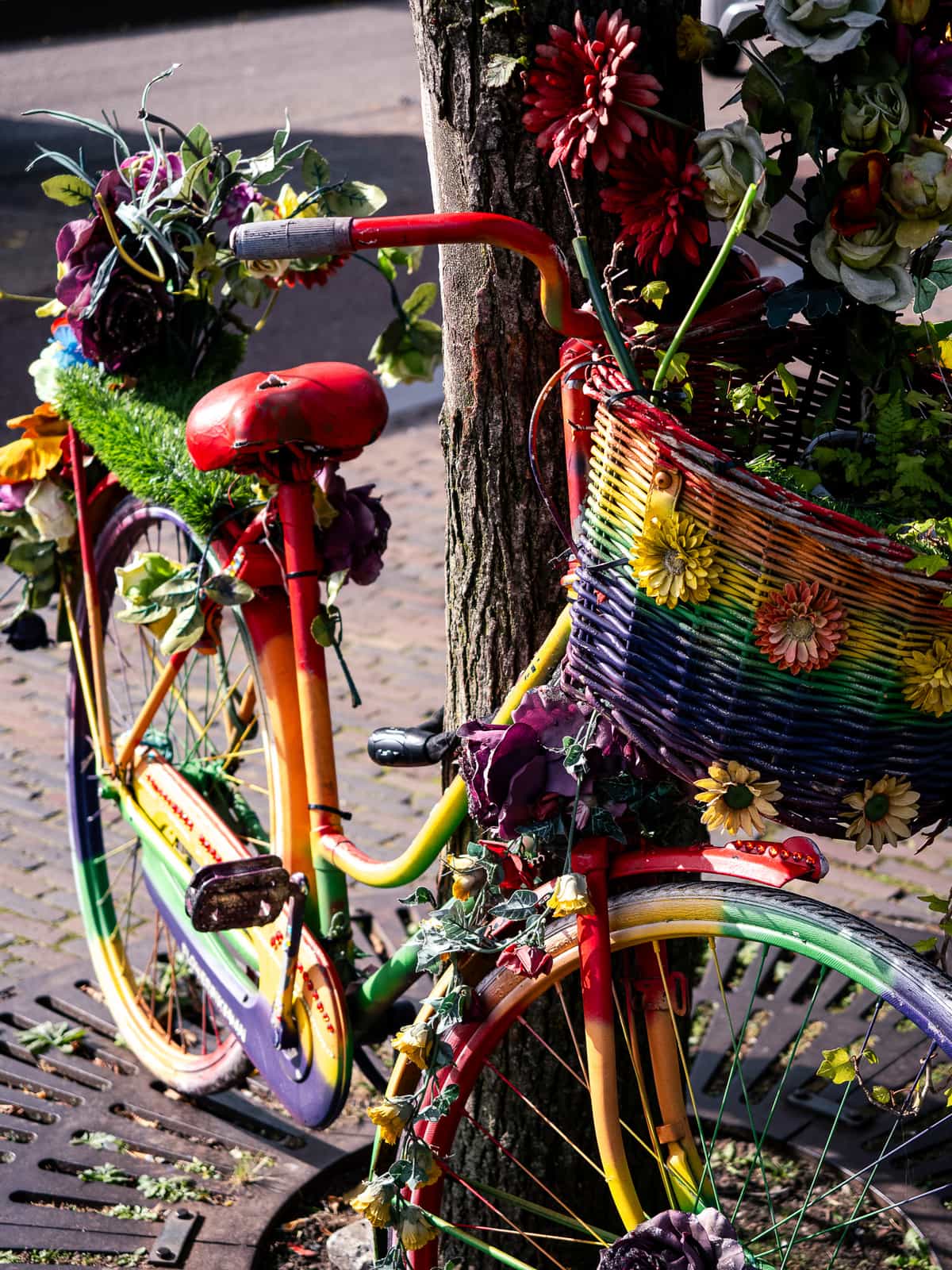 A bicycle that is covered in rainbow paint and flowers leans on a tree