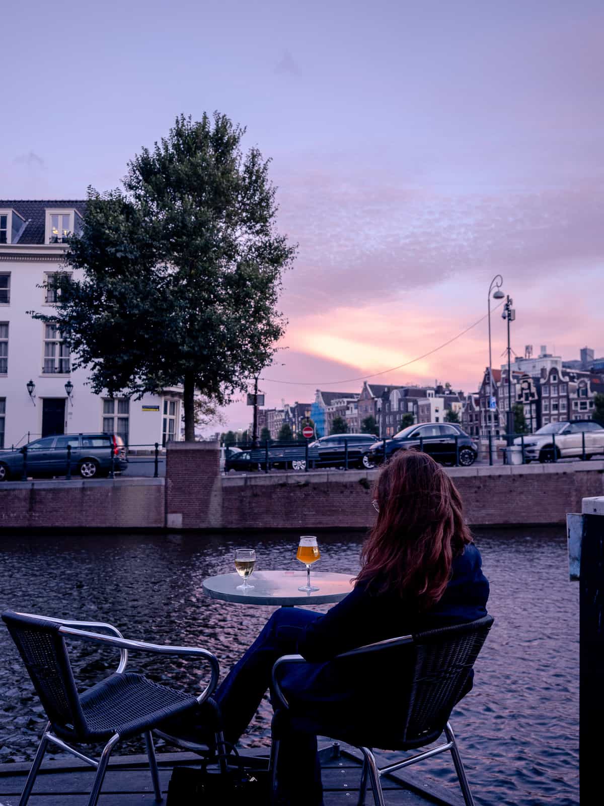 A woman sits at a cafe table with a beer, on a waterfront Amsterdam canal at sunset