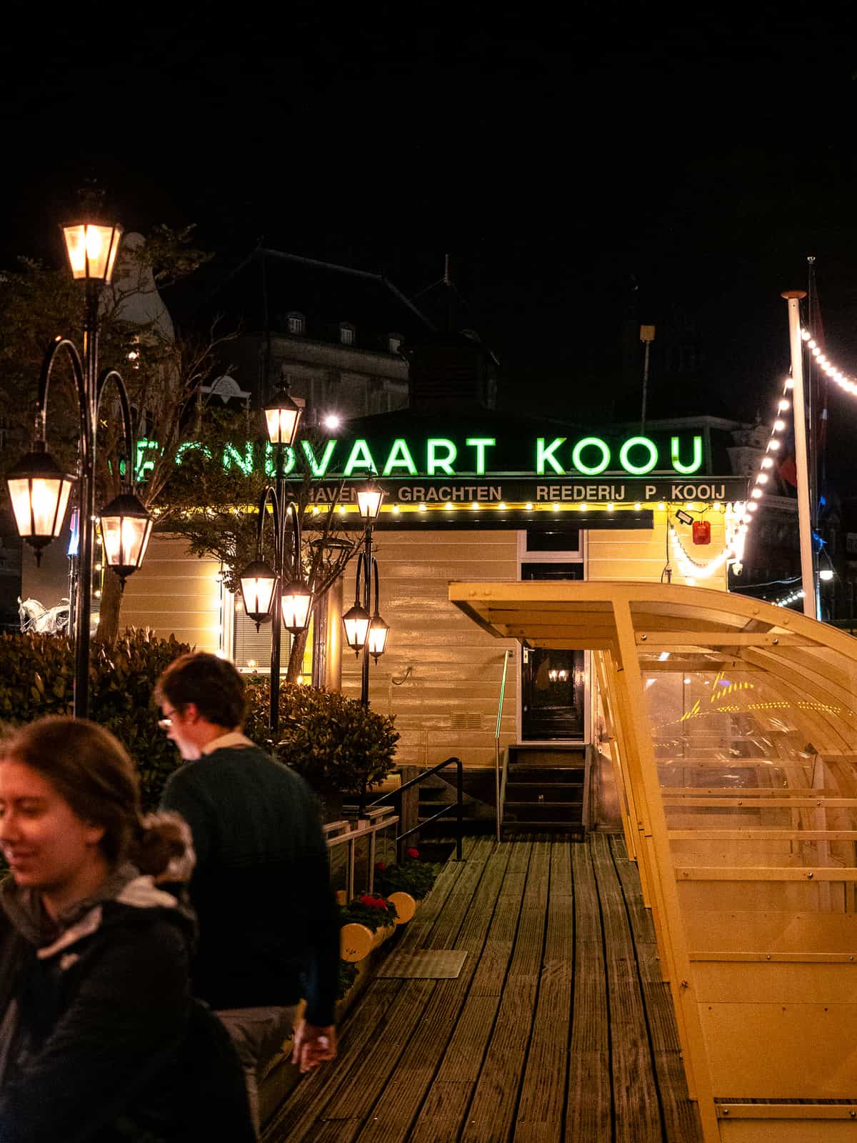 A sign reading "Fondvaart Koou" glows in green neon at night, as people walk by