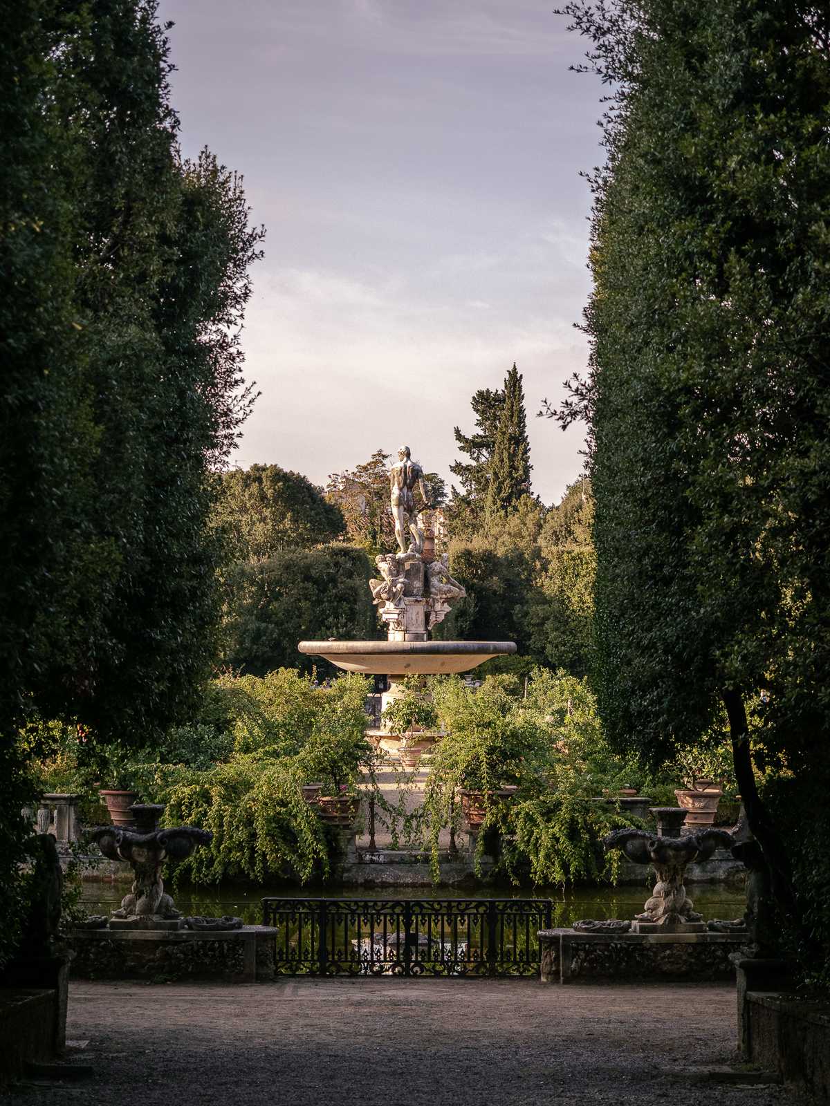 A fountain with a male statues on top catches sunset light, looking from between two dark hedges