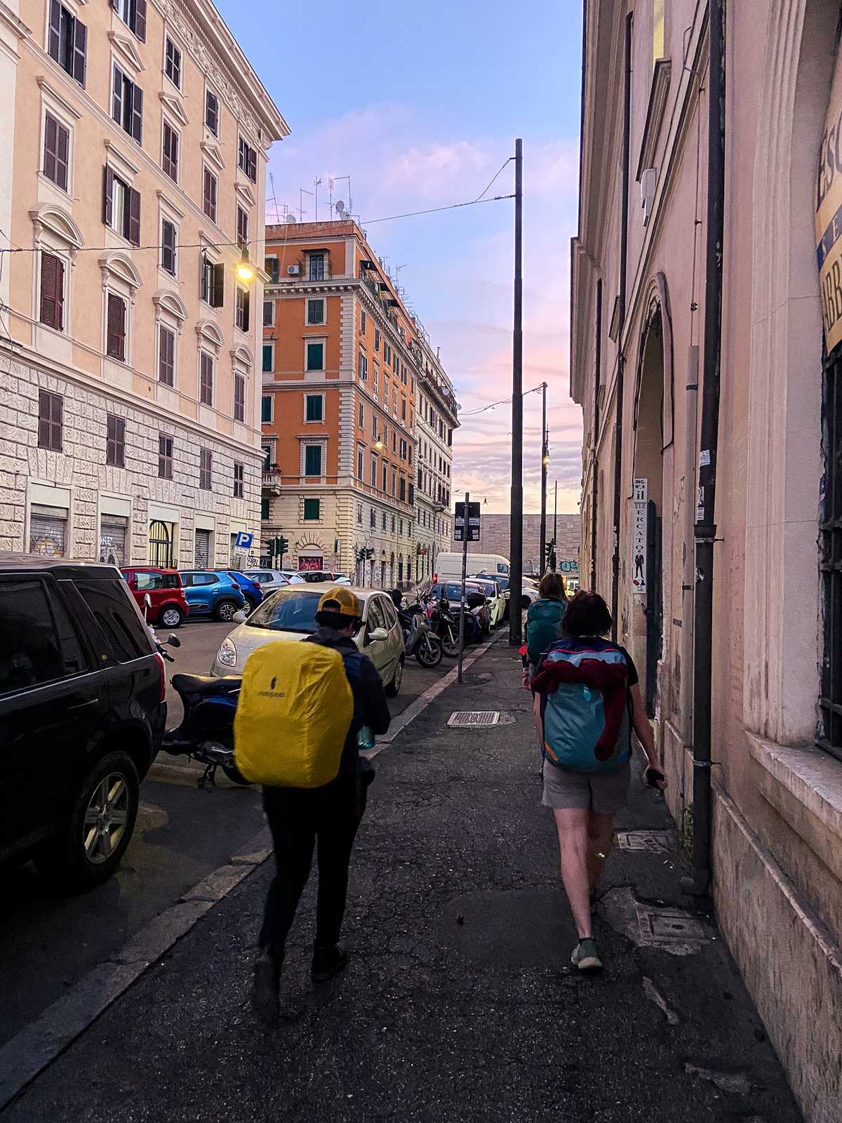 Backpackers walk down a street toward the ocean at sunset, in Naples, Italy