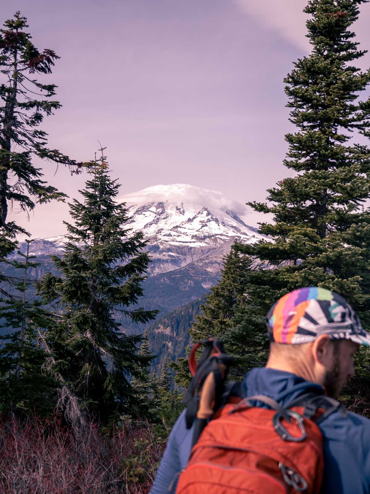 Mount Rainier in the distance with a cloud on top of it, surrounded by pine trees, and the blurry outline of a person in the foreground.