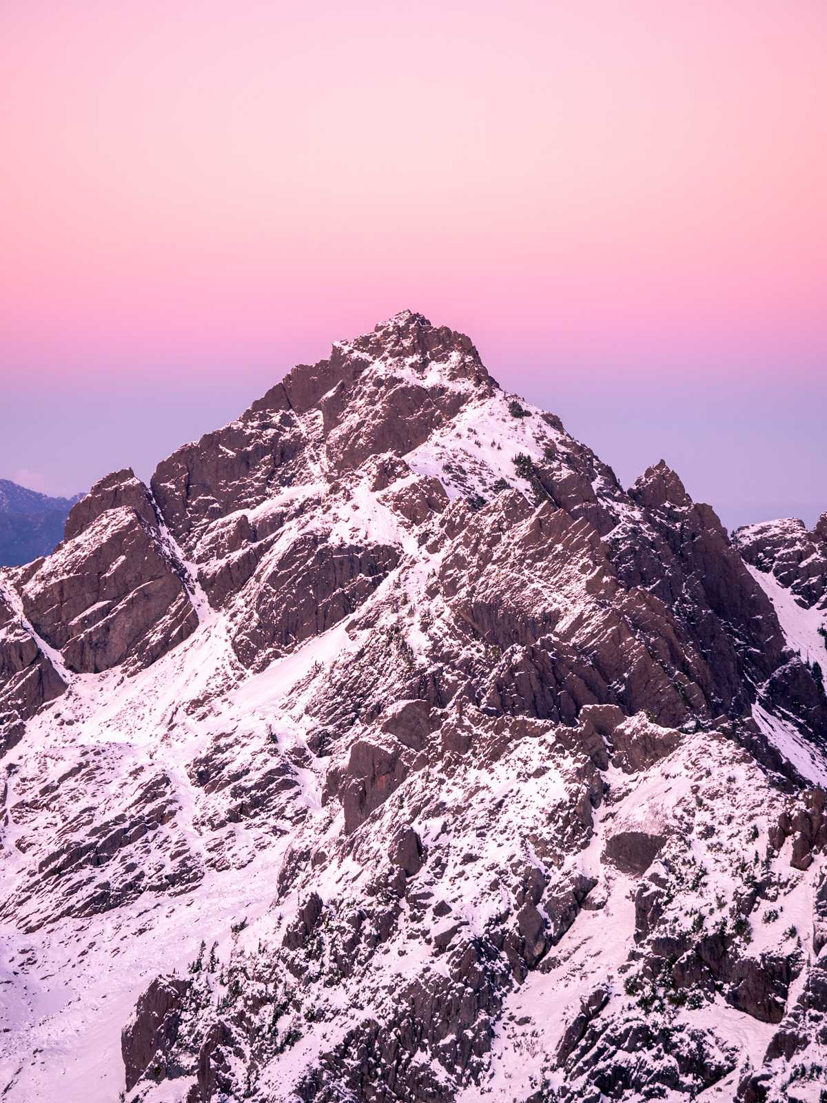 Sunset gradient of pink to purple lights, the sky, but behind a triangular shaped mountain ridge covered in snow poking into the sky