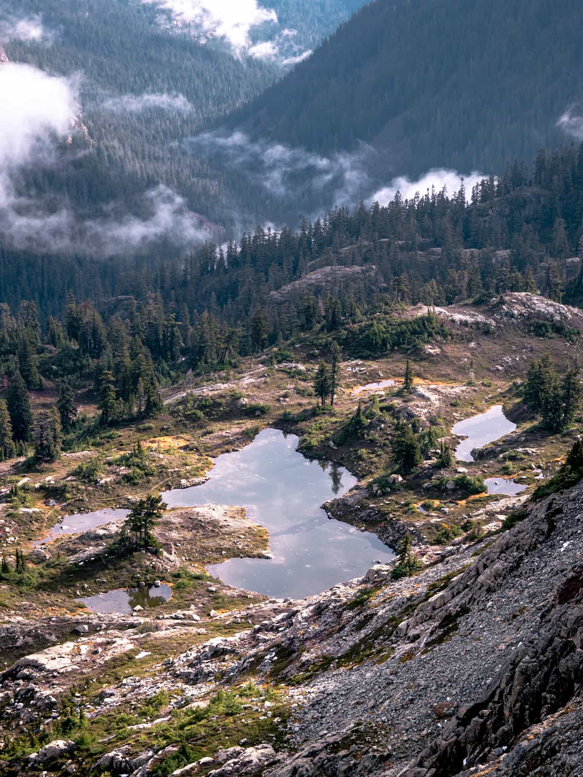 Alpine ponds reflect cloudy sky with clouds, drifting through wooded valleys in the distance