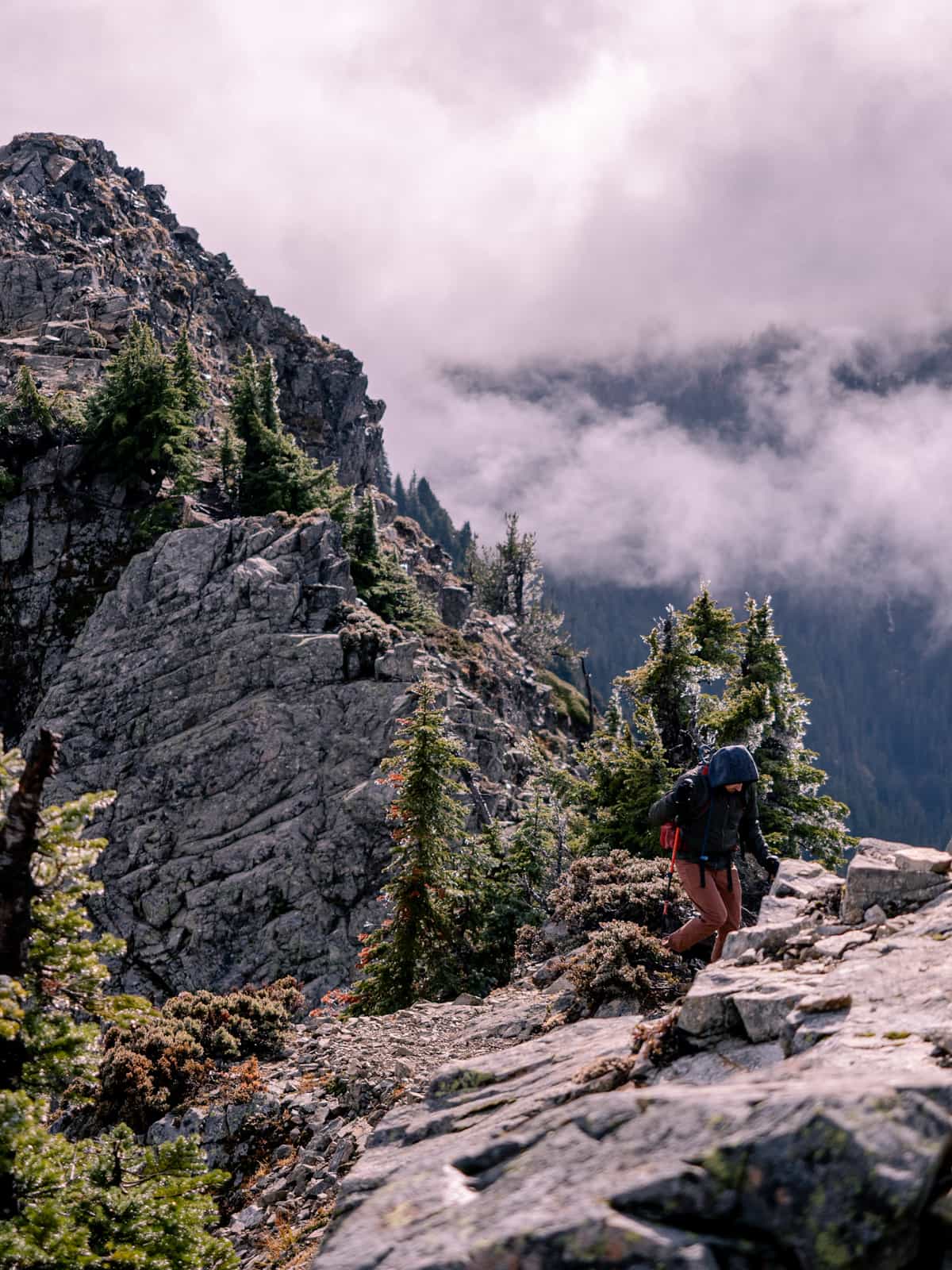 A person hikes narrow exposed ridge line which trees and sharp rocks