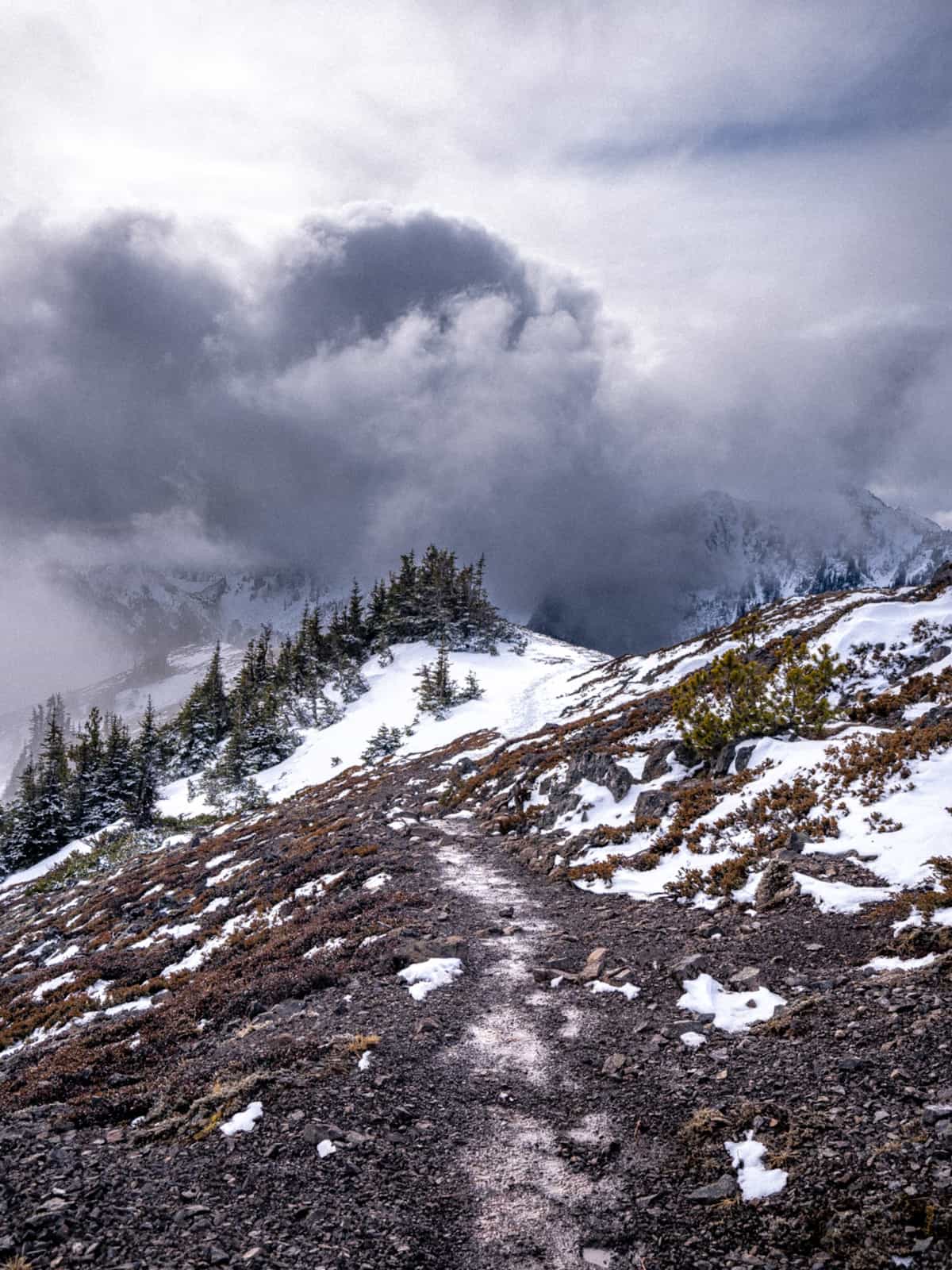 A trail goes down a snowy hill towards dramatic looking clouds