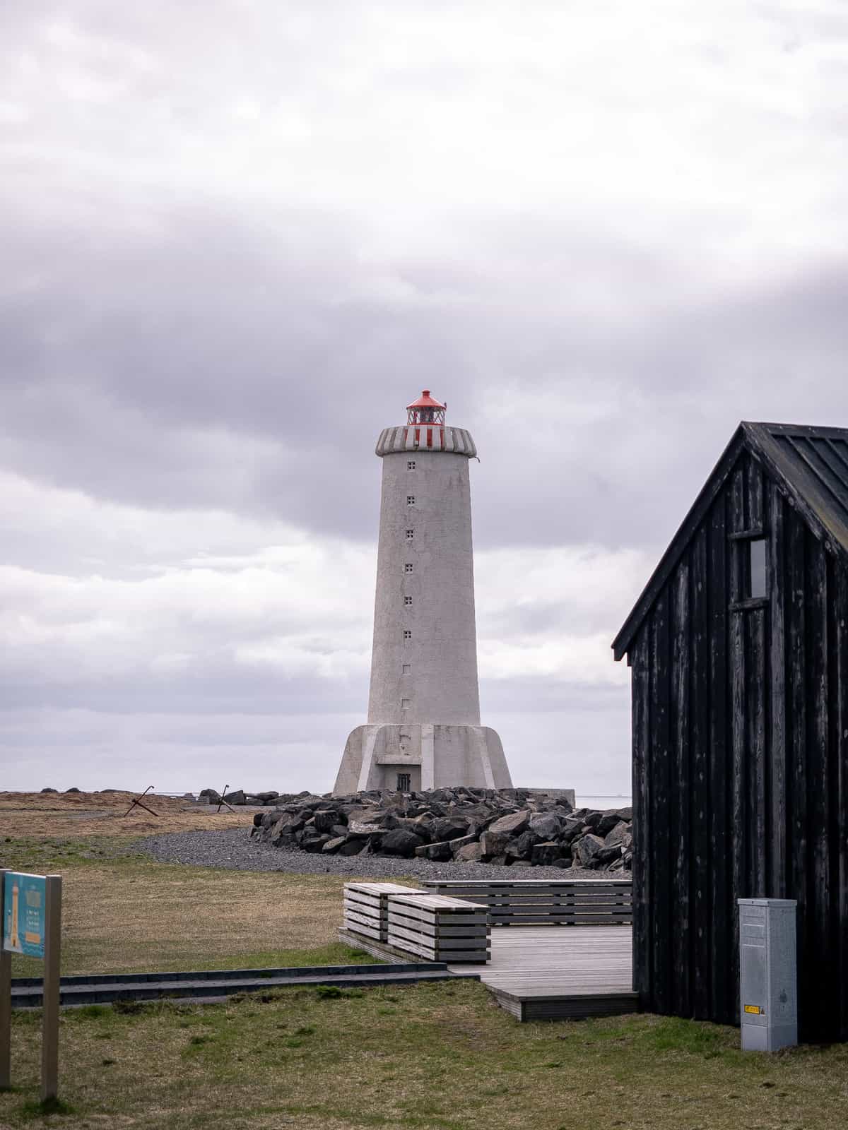 A lighthouse extends into a cloudy sky, with a dark black wood panelled building in the foreground.