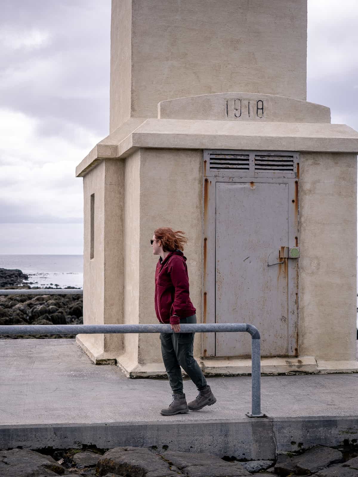 A woman stands at the base of a white lighthouse looking out over the water, hair blowing in the wind.