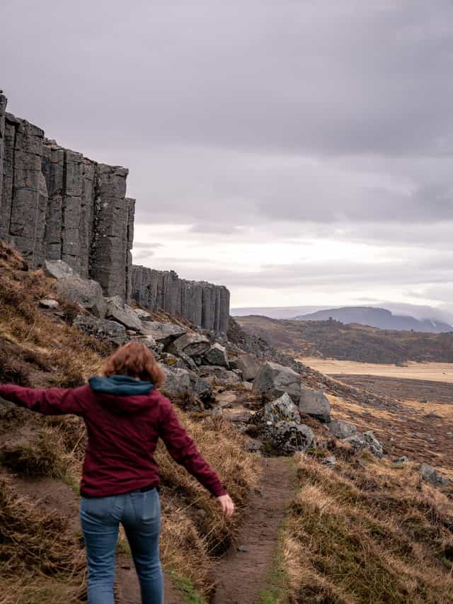 A woman walks, arms waving to the sides, below a cliff of hexagonally shaped rocks.