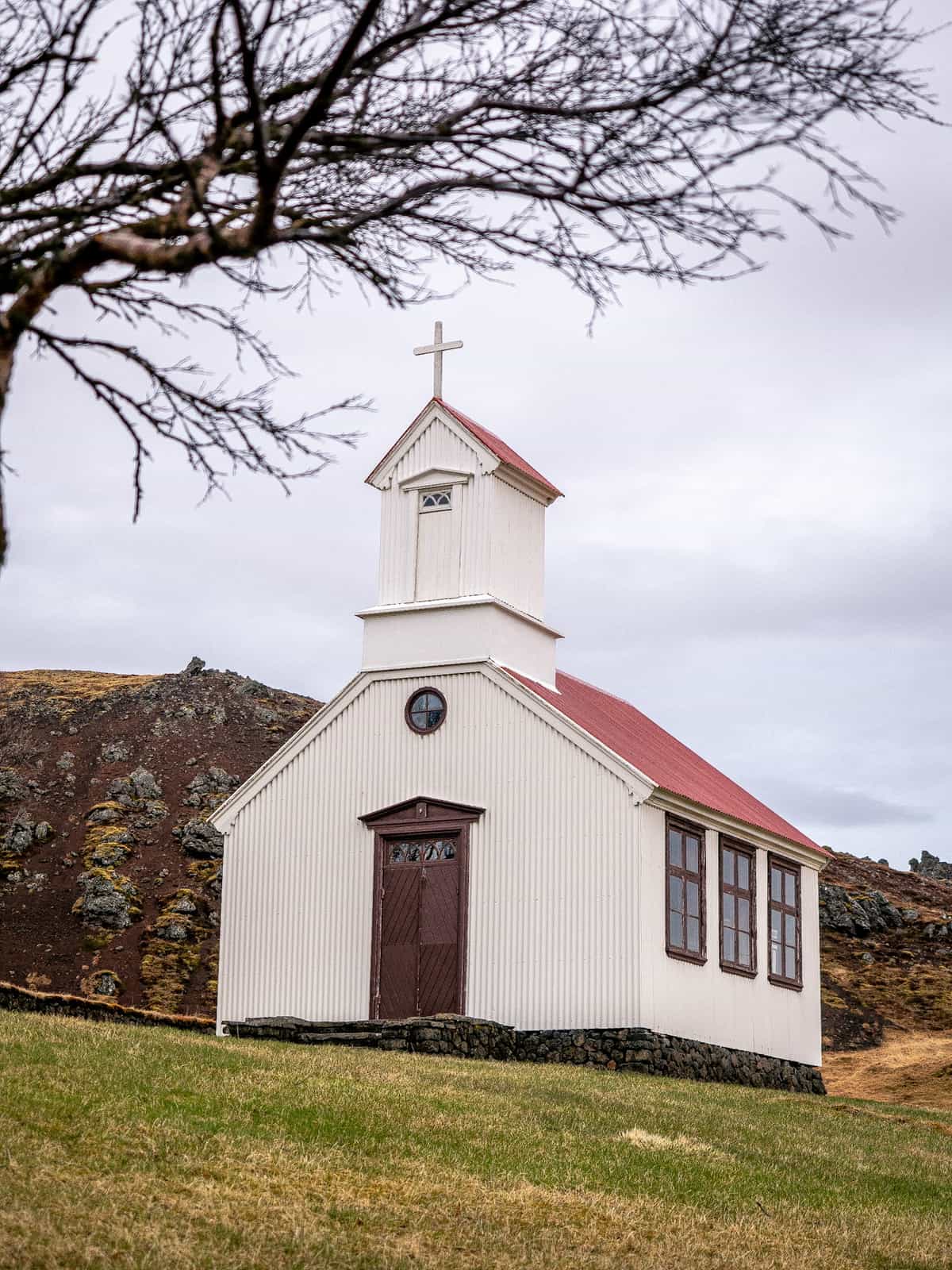 A write and brass colored church sits below a dirt-covered hillside, with blurry branches in the foreground.