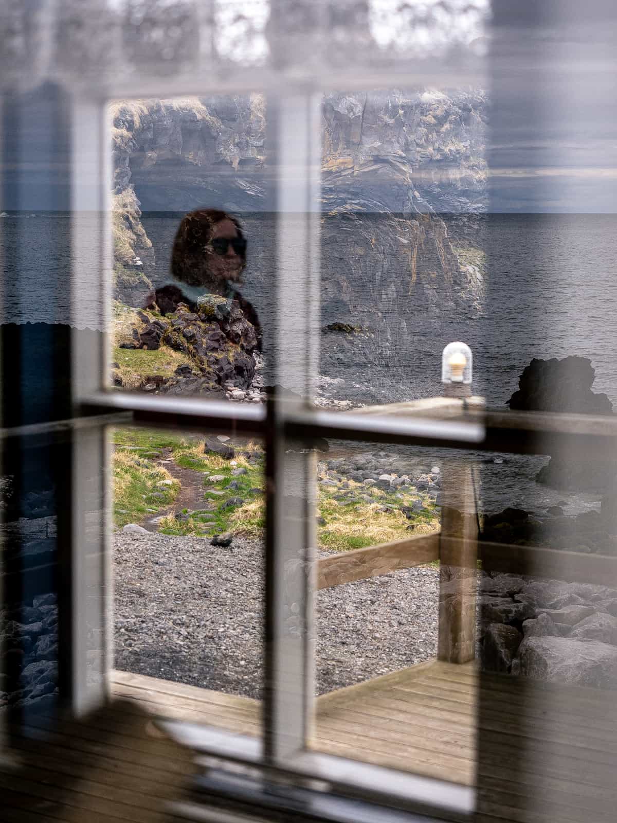 Looking through two windows, a person reflects in the first, with jagged rocks seen through the second