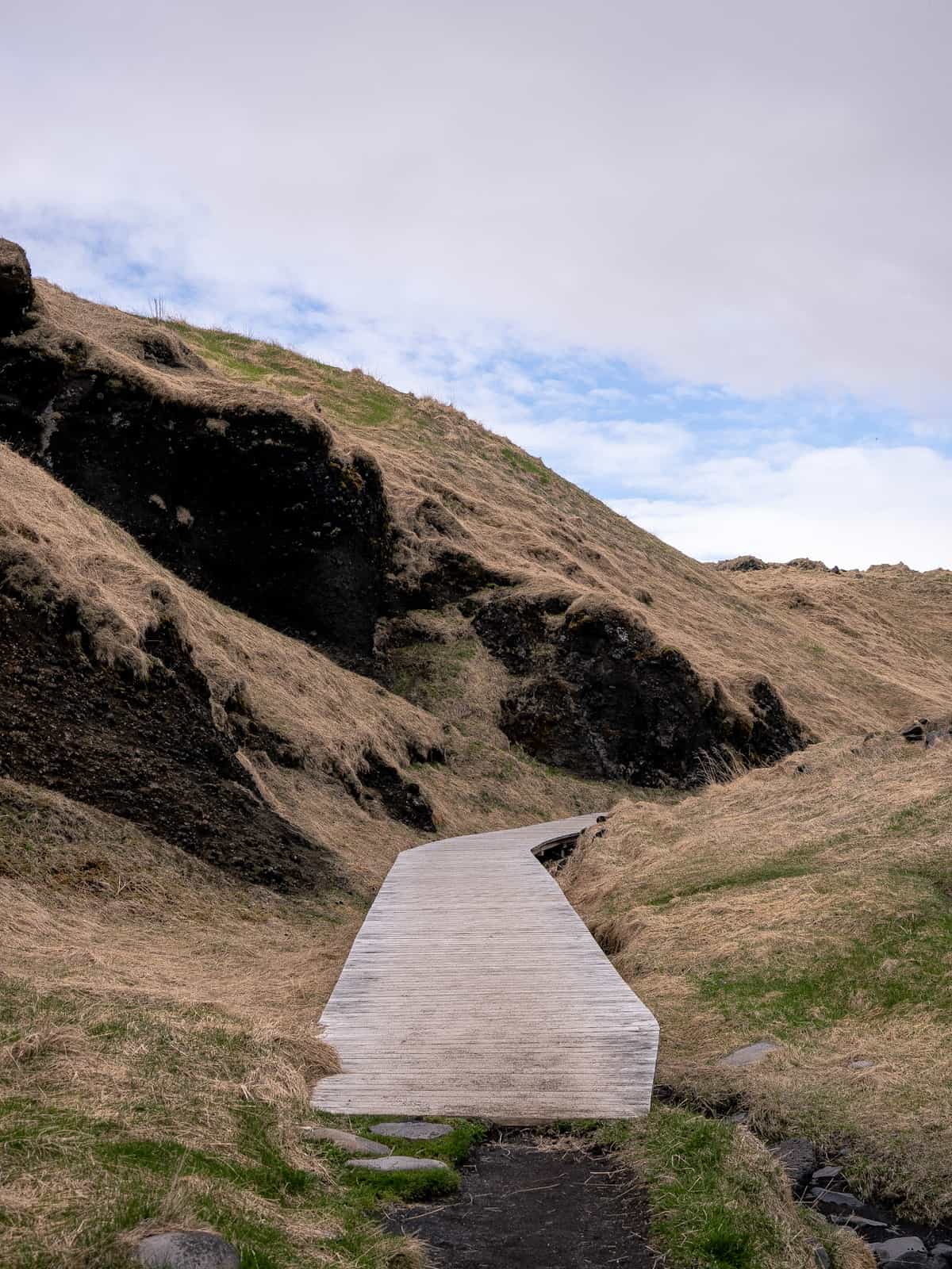 A wooden pathway extends between two grass hills, curving around an unseen corner