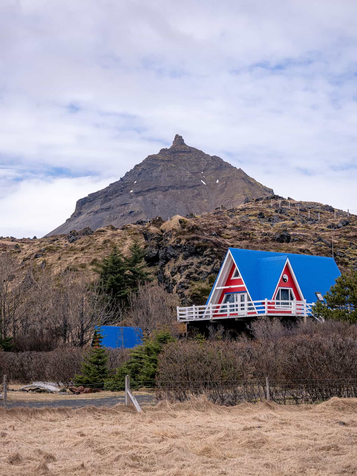 A bright red and blue house sits below a hillside, with a tall mountain in the background