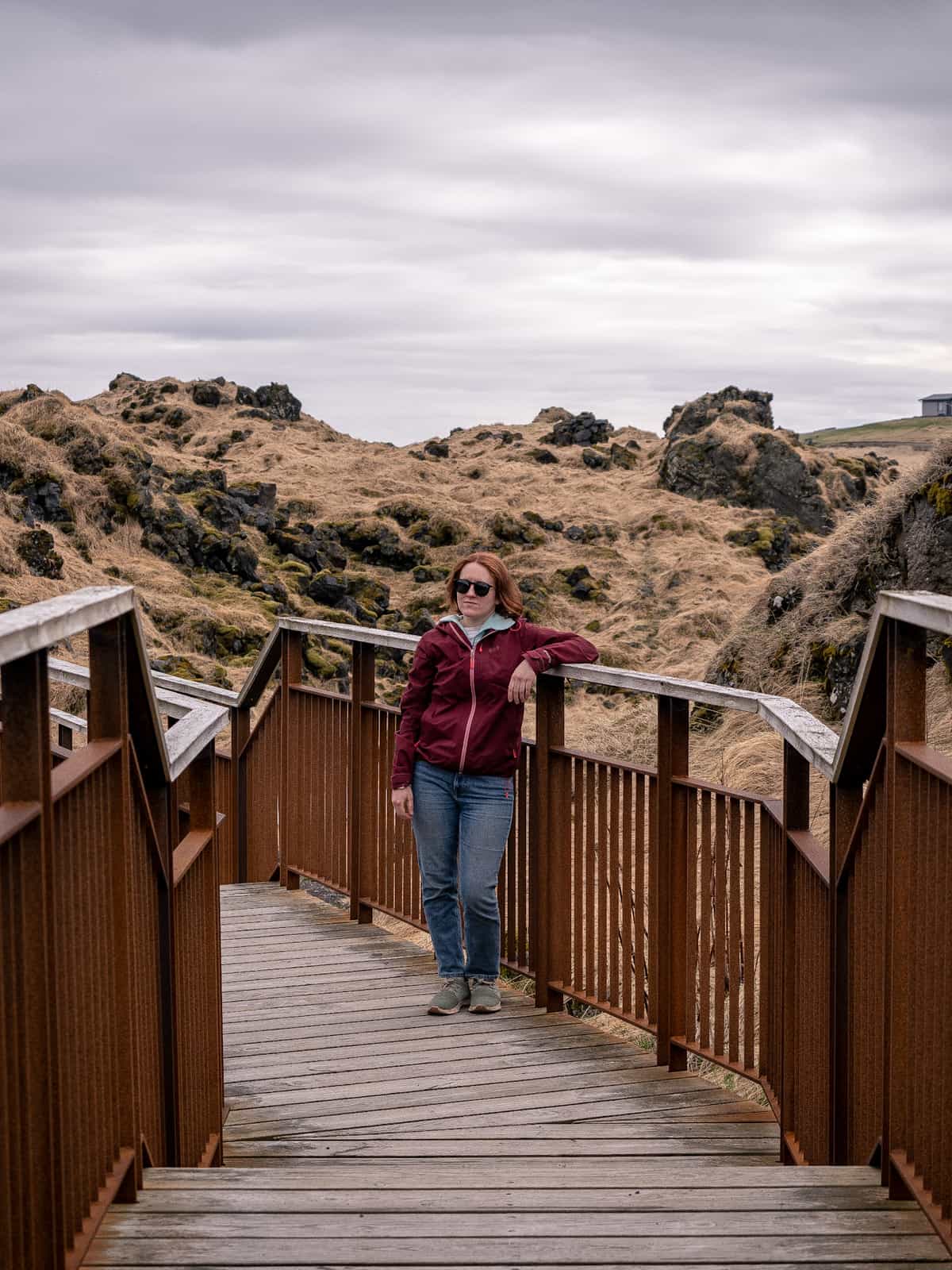 A woman leans against the brown railings of a wooden pathway between two grassy hills