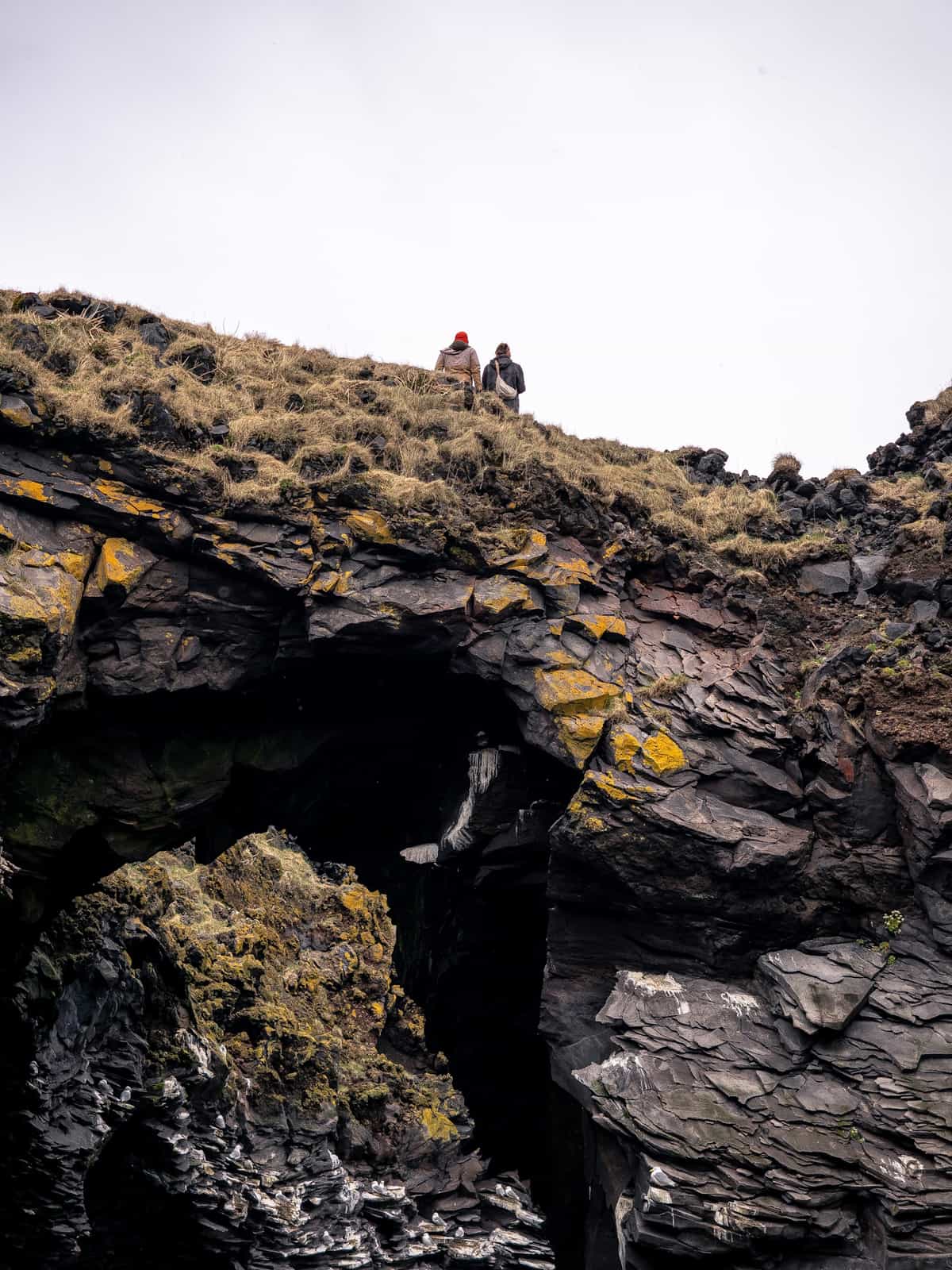 Two people walk on top of a stone archway covered in grass and moss