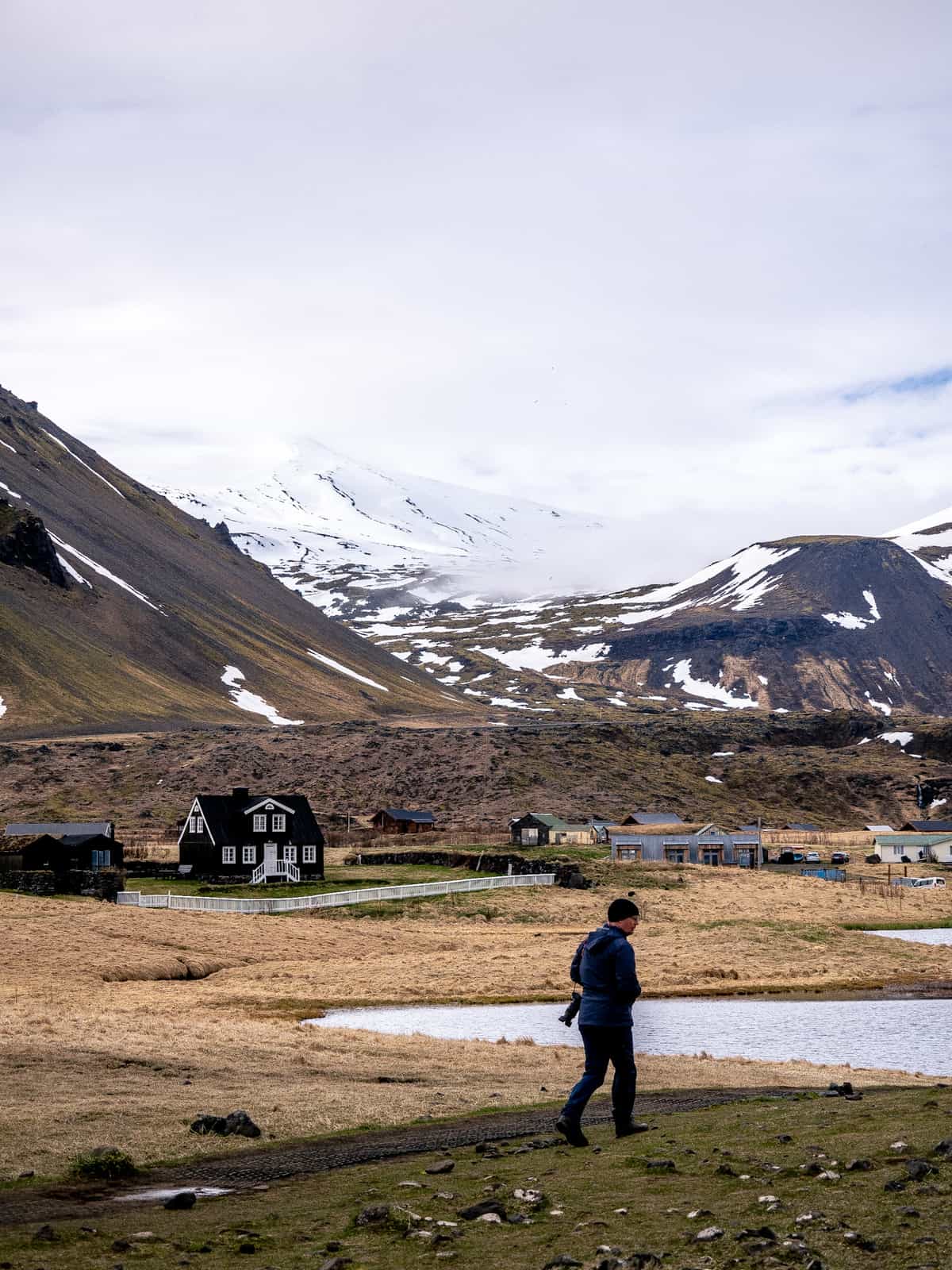 A photographer walks along a pathway, with houses in the middle distance and snowy, white-capped mountains in the far distance