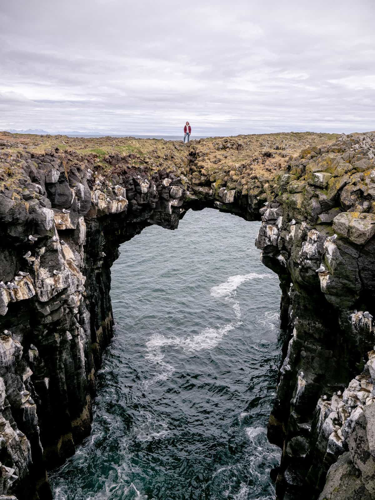 A woman stands on top of a stone archway, extending over the ocean