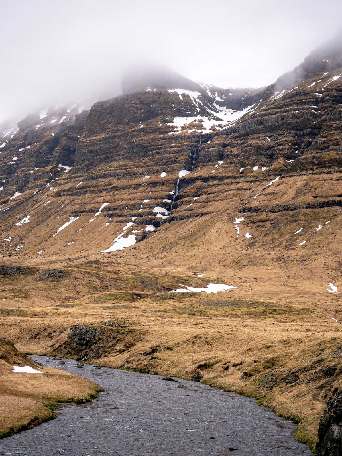 A waterfall moves down repetitive ridges of a golden mountain side, with a river in the foreground