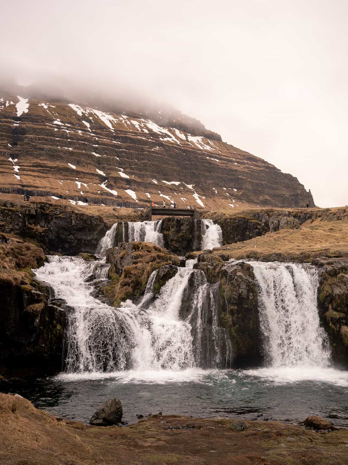 A cascade of waterfalls flows beneath a footbridge, with large hills in the distance