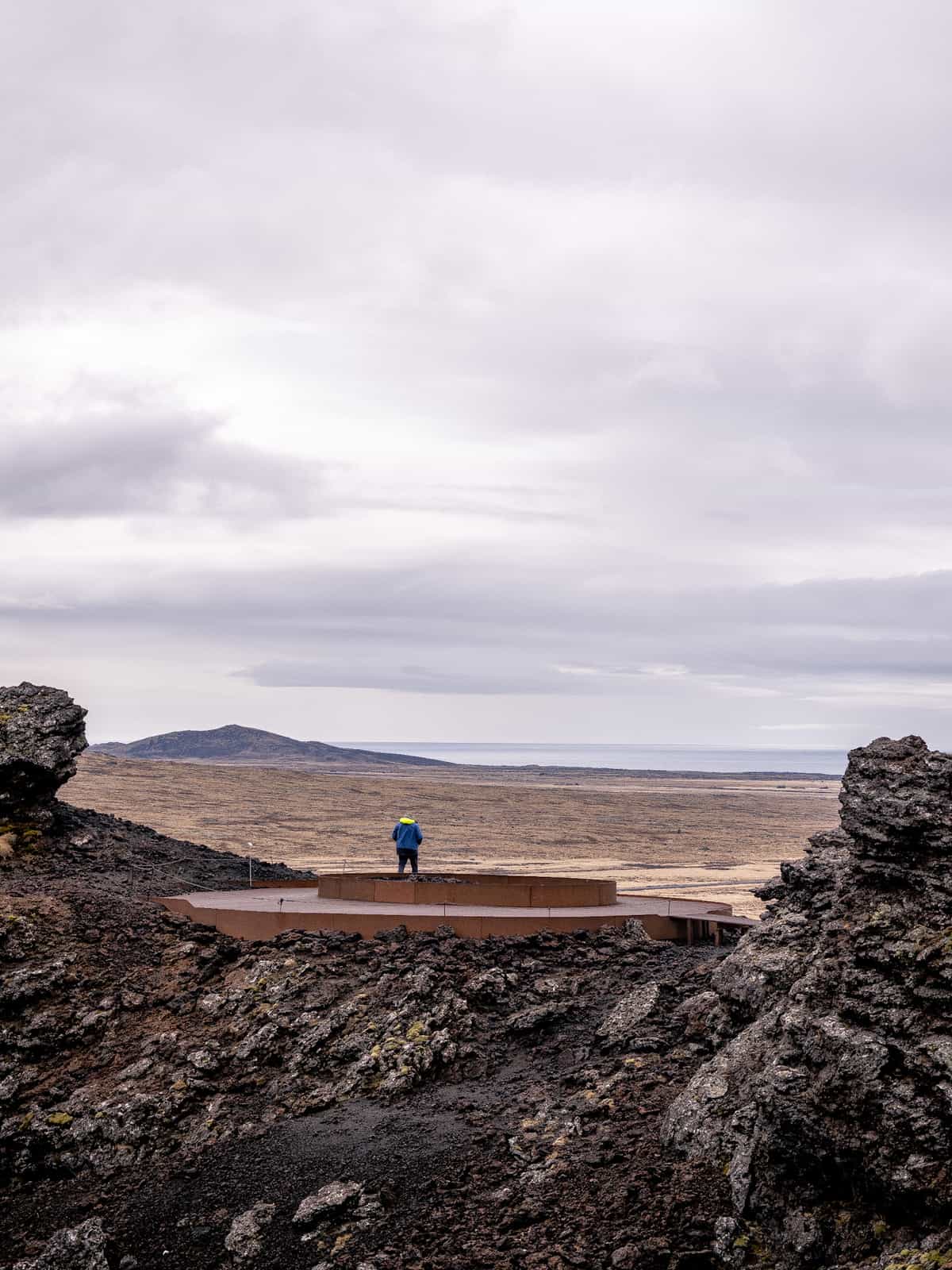 A person stands on metal grating on top of a dark hillside, with plains and ocean visible in the distance