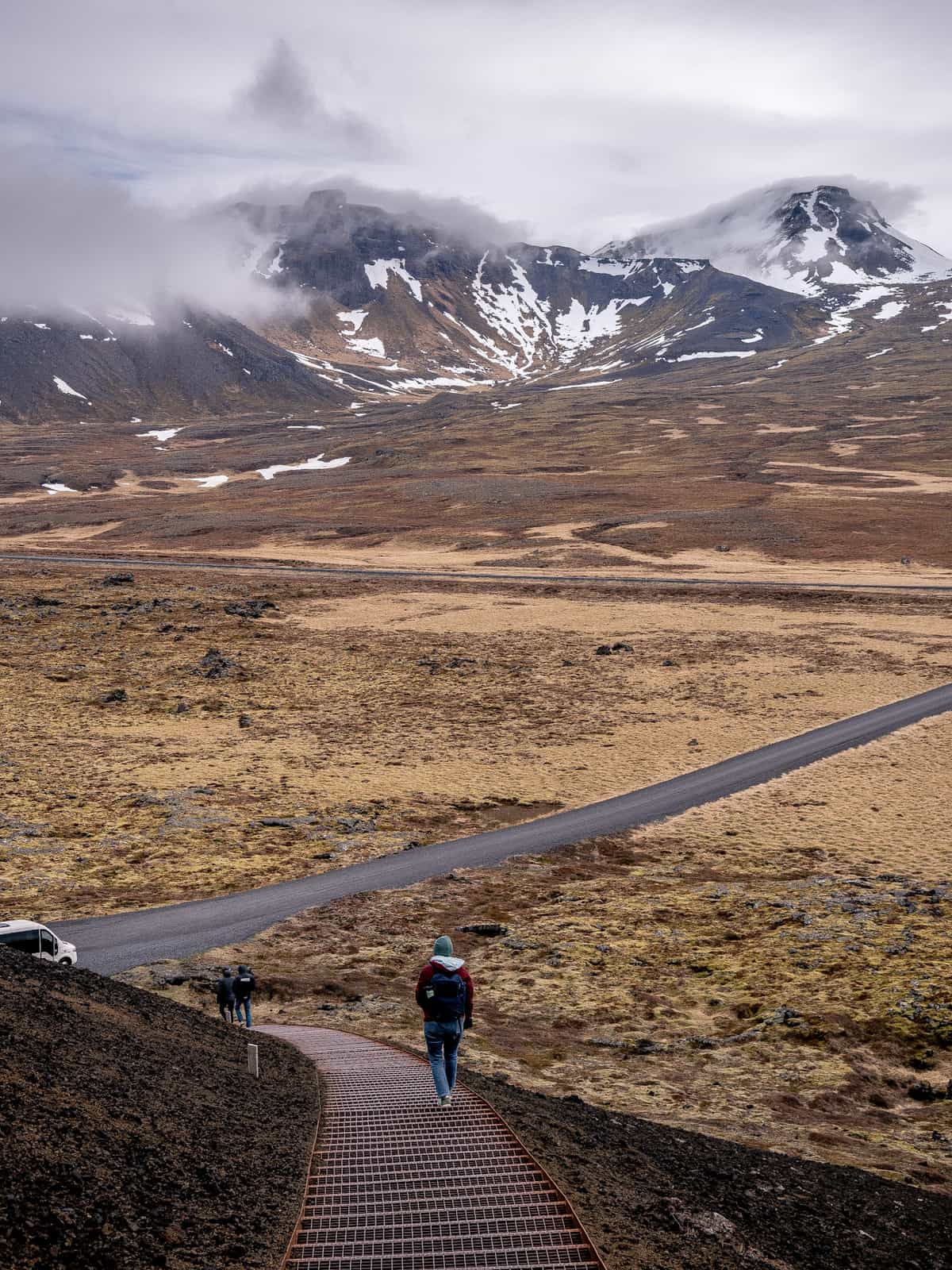 A person in a red jacket walks down a curved metal walkway, with snowy mountain visible in the distance