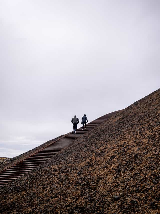 Two people walk up a metal staircase along a brown rubble hillside