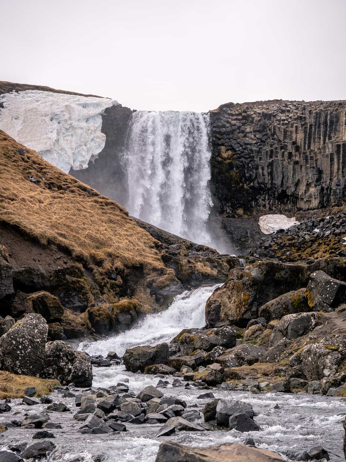 A waterfall cascades down a cliff, with hexagonal stone columns and snow next to it