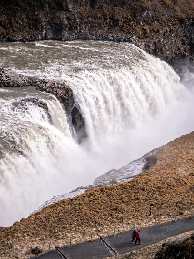 Two people walk down a pathway, across from a very large and wide waterfall