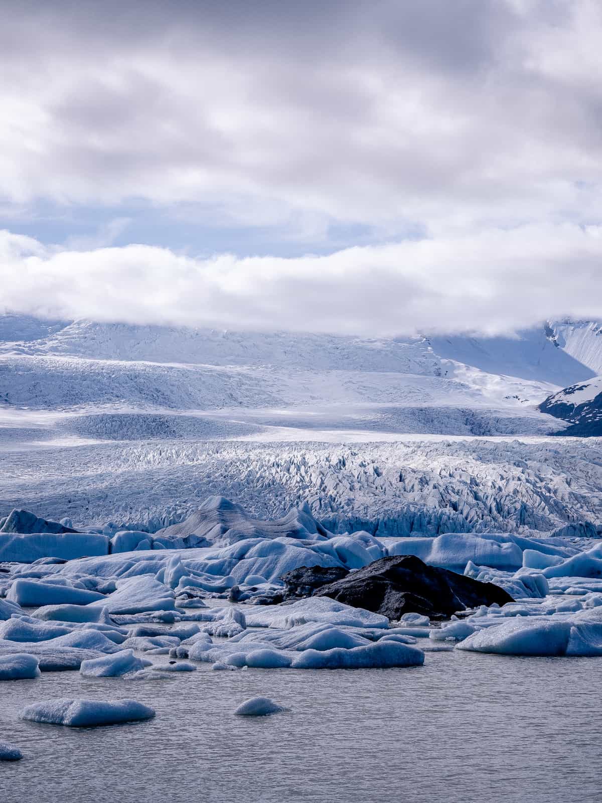 Under mixed clouds, a glacier rolls down a hillside, with a glacier lagoon in the foreground