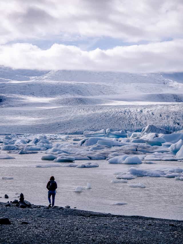 Under mixed clouds, a glacier rolls down a hillside, with a glacier lagoon in the foreground and a woman walking along a rocky beach