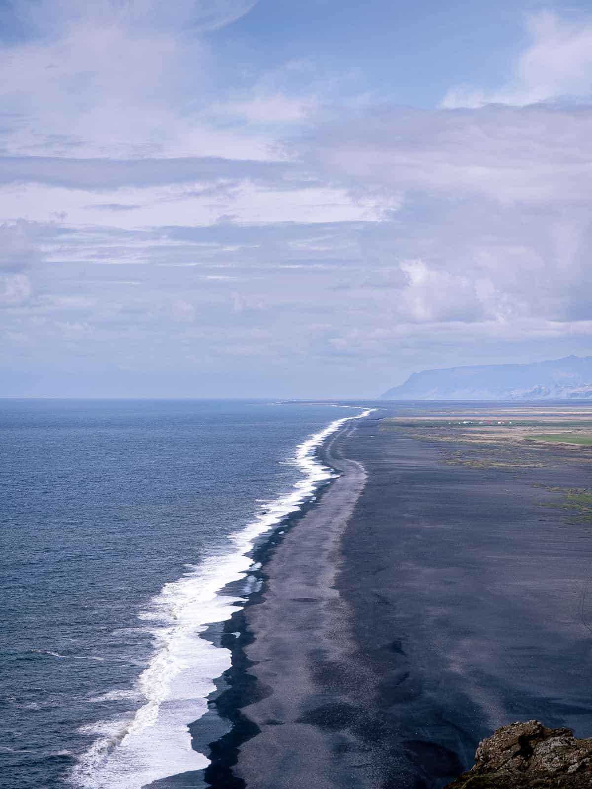 White ocean waves crash on a black beach, with mountains in the distance