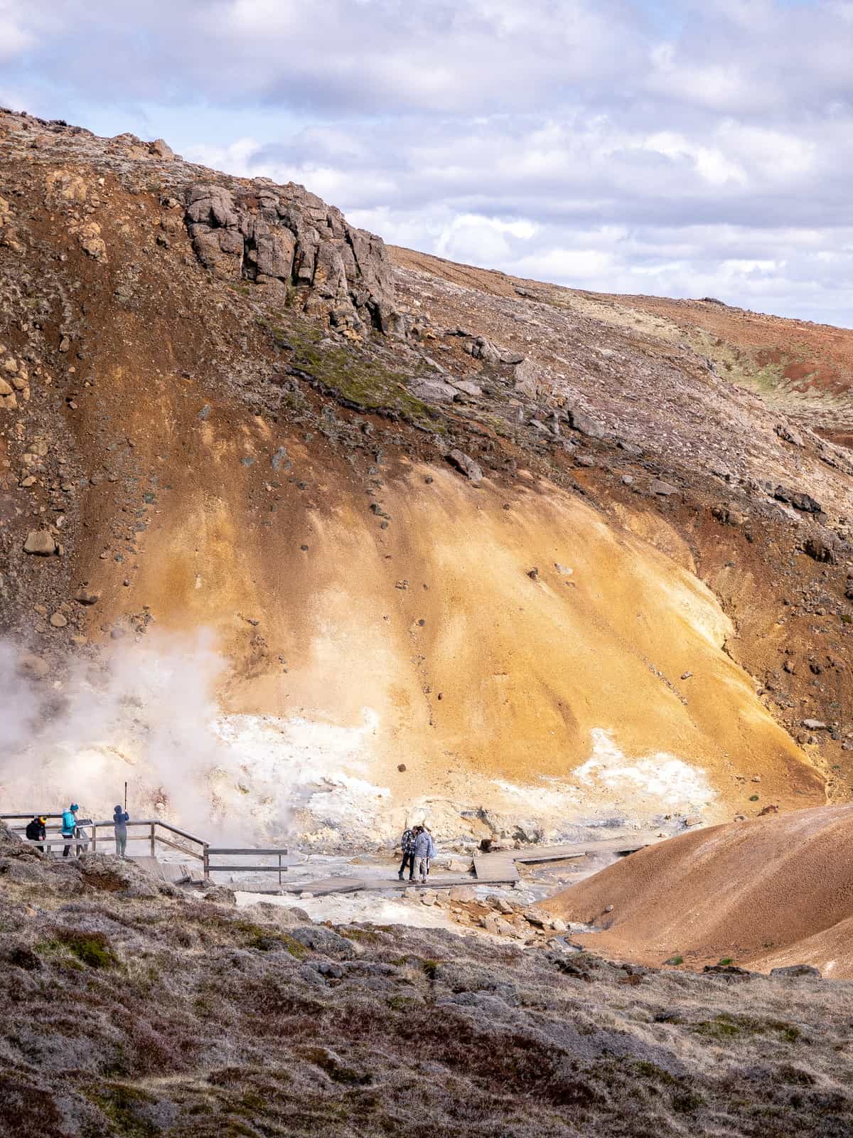 People walk along a pathway cutting across geothermal pools and mud pits