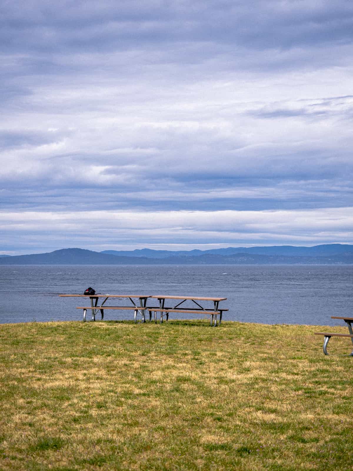 Picnic benches sit on a grassy knoll looking out over ocean water with mountains, visible in the far distance