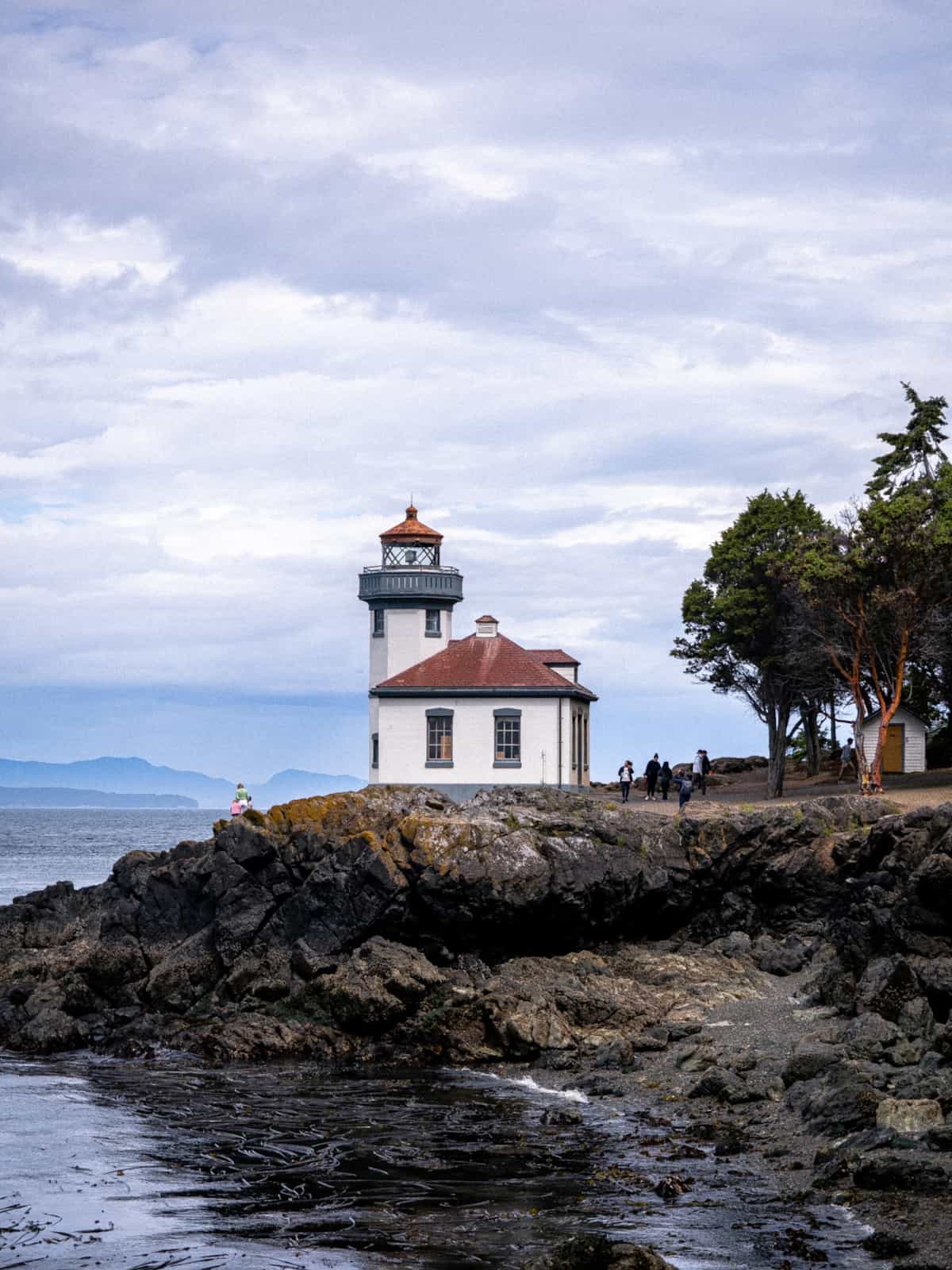 A lighthouse sits on rocks with trees next to it and people walking around